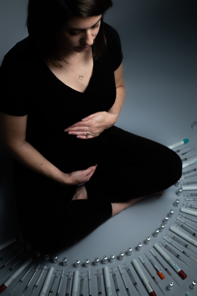 Kristen seated cross-legged on a grey backdrop, looking down, arms cradling her pregnant belly. Laid out in front of her in a decorative semicircle are all her needles and vials from her IVF treatments. Image is taken from a 45 degree angle to Kristen and slightly from above.