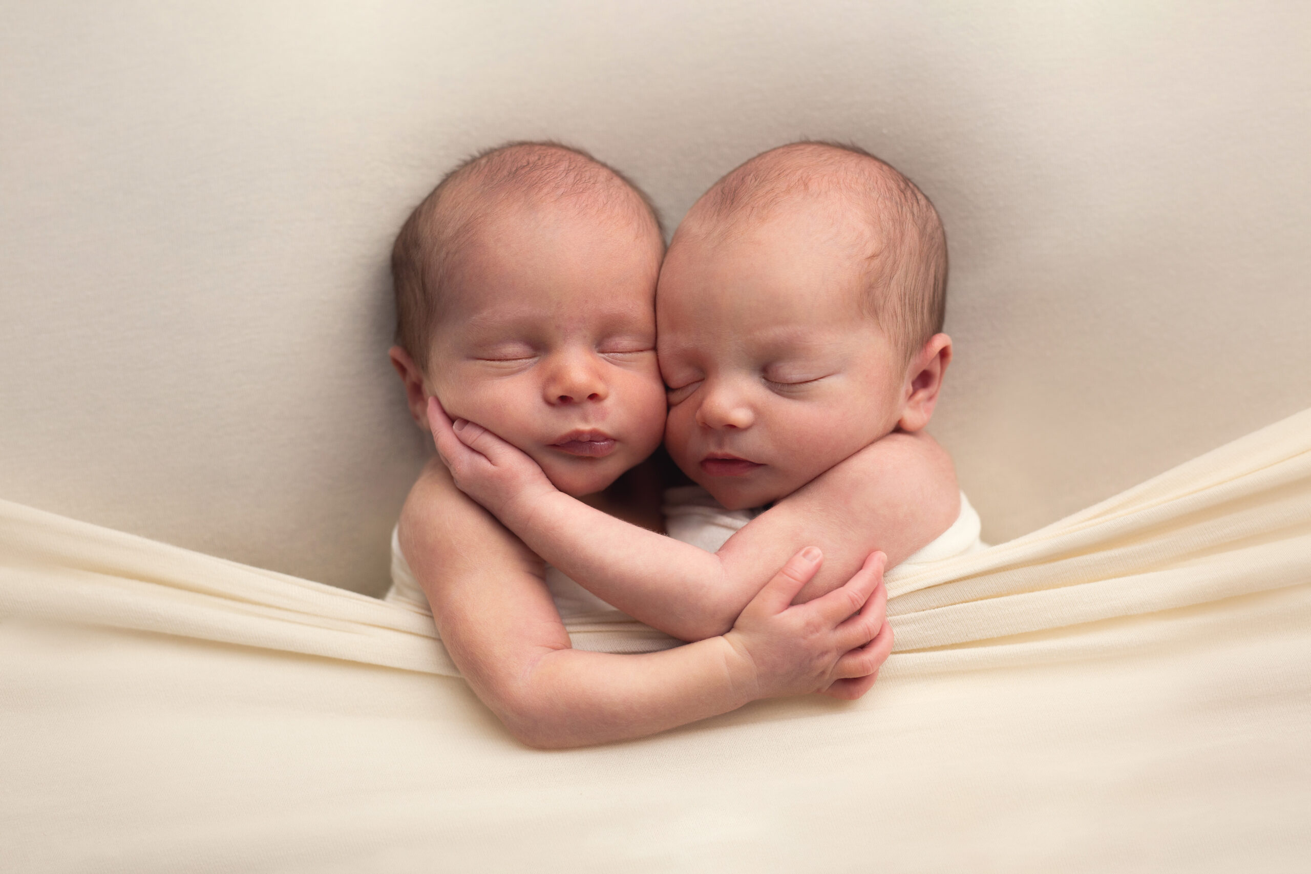 Twin newborns tucked in on ivory coloured sheets snuggling together with their arms around each other