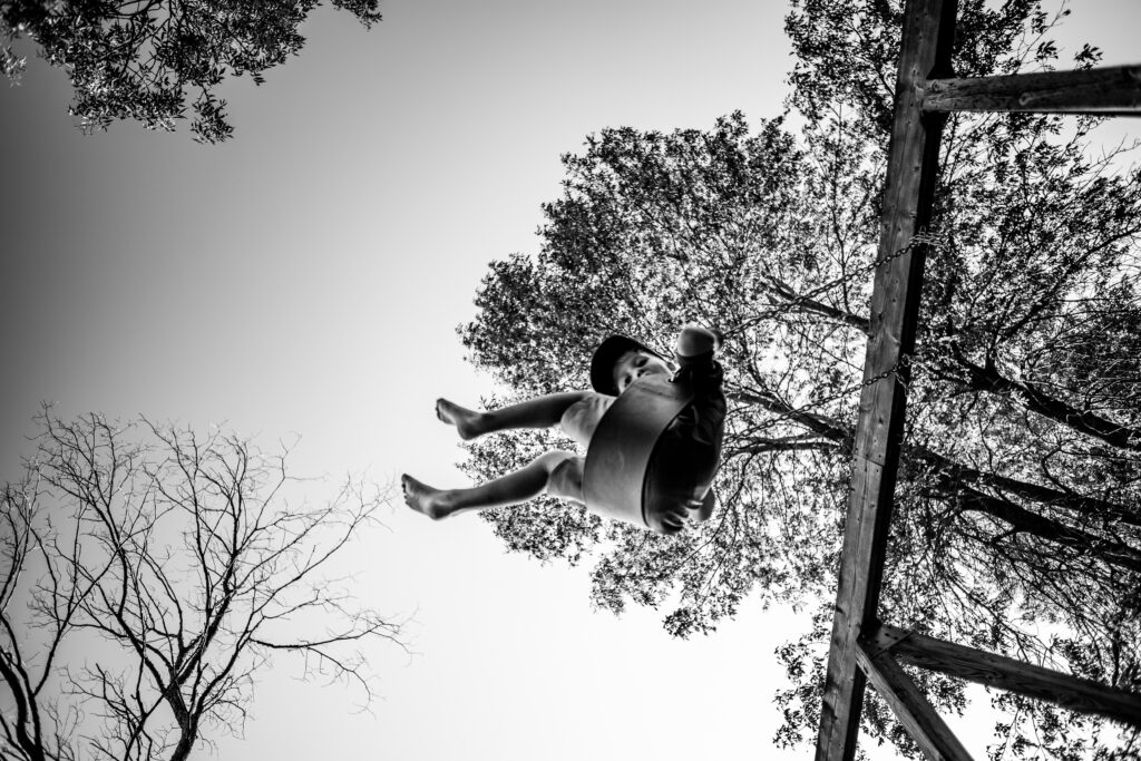Black and white image of seven year old boy swinging on a swing, taken from low angle.