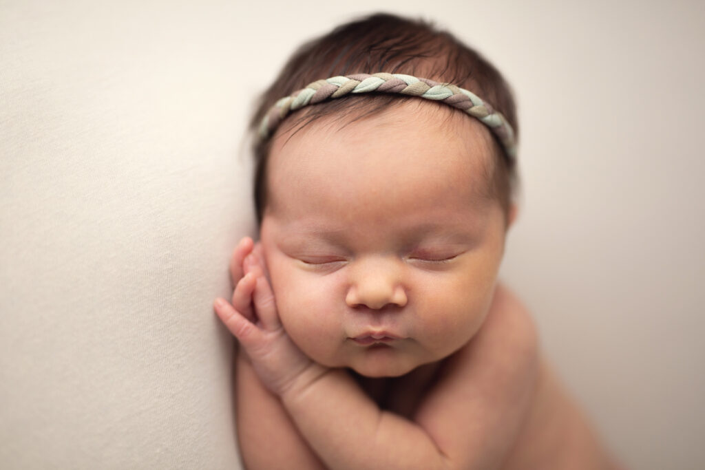 Sweet portrait of a sleeping newborn baby girl wearing a soft braided headband, nestled peacefully on a textured blanket, showcasing the curled-up, dreamy newborn pose best captured in the first few weeks after birth.