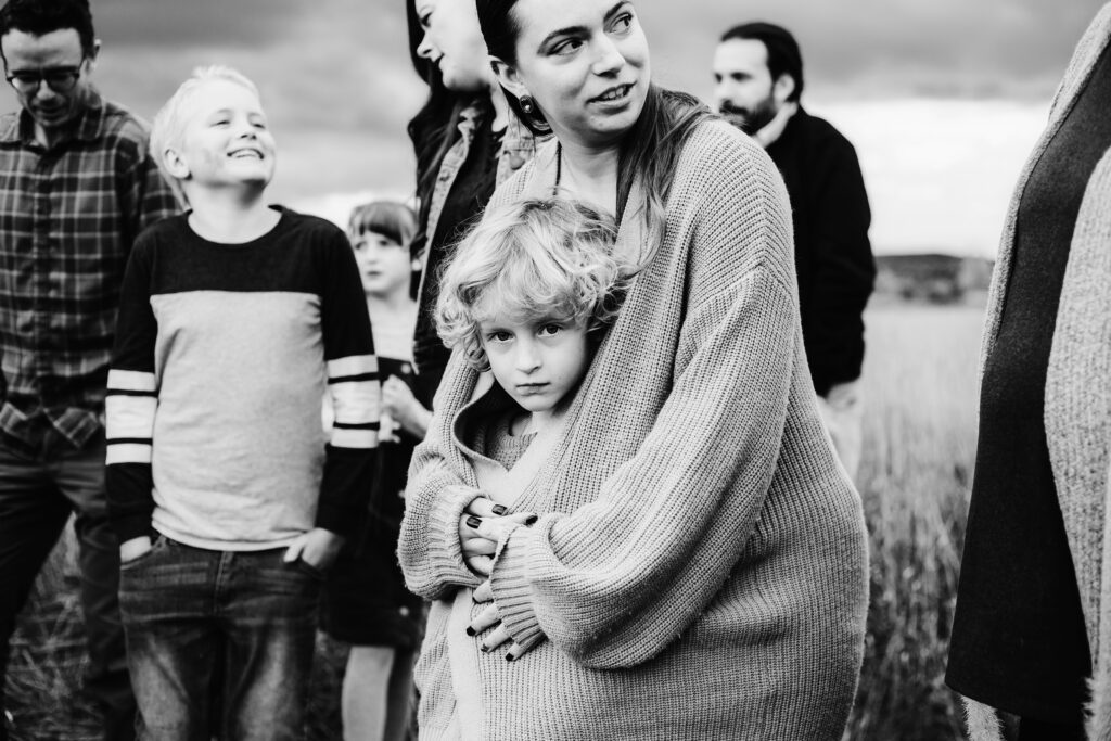 Black and white image of child wrapped in her aunt's sweater looking at the camera while extended family members interact in the background at a Collingwood extended family session.